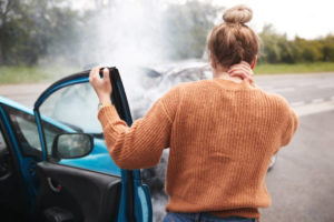 A woman wearing a brown sweater steps out of the passenger seat to look at the car crash in front of her.